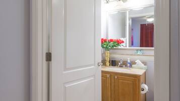 A bathroom with a white door, wooden cabinets, a sink with a mirror above, and a vase with red flowers.