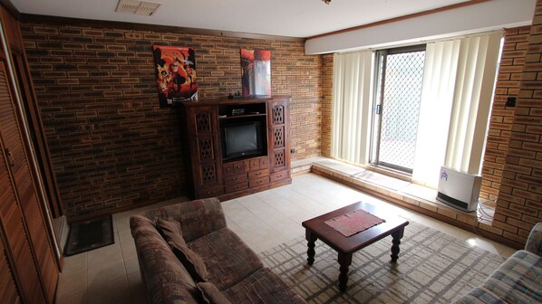 A living room with a brick wall, a sofa, a coffee table, a TV stand, and a large window with blinds.