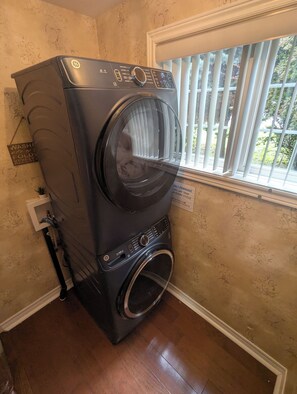 A front-loading washer and dryer unit with a control panel, a wall-mounted sign, and a window with blinds. - River Views | Pool Table | EV Charger | Hiking Trails | Near Niagara Falls (St. Catharines)