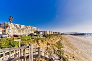 A coastal hotel with a wooden staircase leading to a beach, a pier in the background, and a clear blue sky.