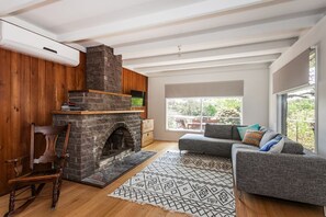 A living room with a stone fireplace, a grey sofa, and a large window offering a view of the outdoors.