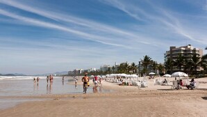 A beach with people walking and sitting under umbrellas, palm trees, and buildings in the background.