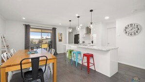 A modern kitchen with a central island, a dining area with a wooden table and chairs, and a large window with curtains. - Seagulls is the ultimate seaside escape! (Lancelin)