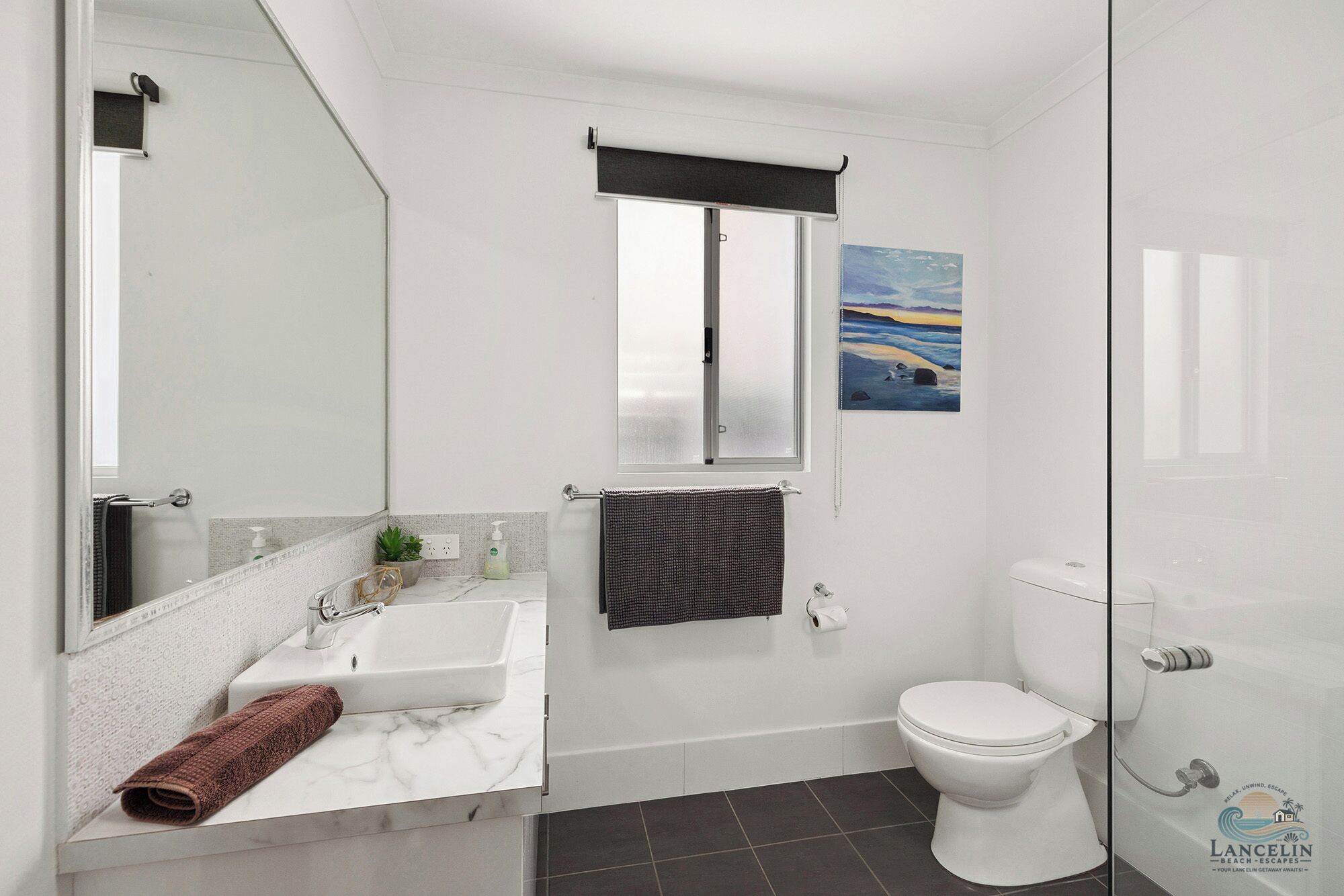 Et moderne badeværelse med en hvid vask, spejl og toilet. Der er et vindue med havudsigt og et billede på væggen.