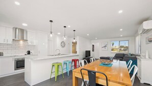 A modern kitchen with white cabinetry, a wooden dining table, colorful stools, and a large window offering a view of the outdoors. - Seagulls is the ultimate seaside escape! (Lancelin)