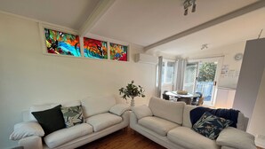 Un salón con un sofá modular blanco, una planta en maceta y un comedor visible a través de una puerta abierta.