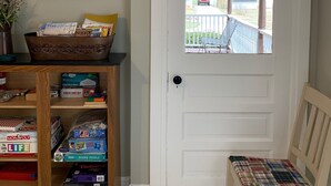 A door with a white valance, a view of a blue house through a window, a chair with a plaid cushion, and a shelf with board games and books. - Idyllic Location - Walk to Town and Beaches (South Haven)