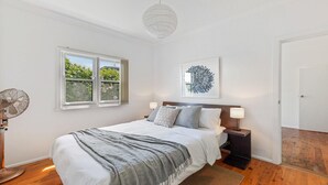 A bedroom with a bed, a bedside table, a lamp, a wall art piece, and a window with a view of greenery.
