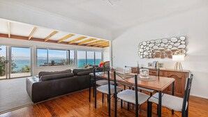 A spacious living area with a large window overlooking the sea, a wooden ceiling, and a dining table with chairs.