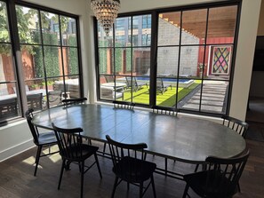 Un espace repas comprenant une grande table ronde et six chaises, de grandes fenêtres donnant sur un jardin avec une piscine et un coin salon extérieur.