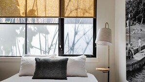 Et soveværelse med seng, puder, lampe, et vindue med skodder og et billede hængt på væggen.