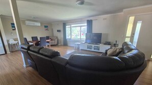A living room with a large leather sofa, a dining area with chairs and a table, a TV on a cabinet, and a window with curtains. - Family friendly Smiths Beach Retreat (Smiths Beach)