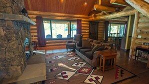 A log cabin interior with a stone fireplace, a large window, a sofa, and a patterned rug. - A Serene Lake Front Cabin Minutes from West Glacier National Park (Columbia Falls)