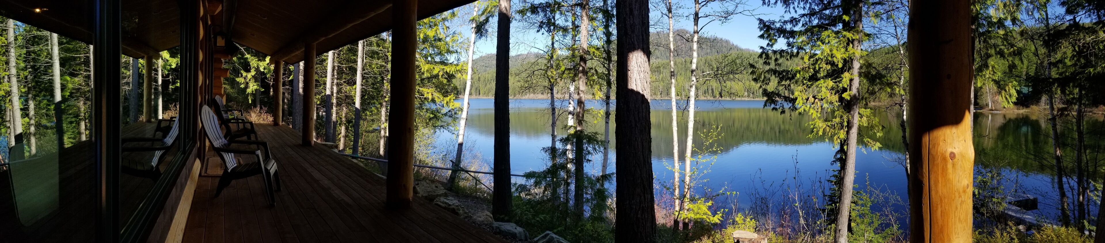 Une terrasse en bois, dotée de chaises, qui offre une vue sur un lac paisible, entouré d’arbres.