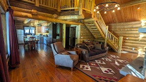 A spacious log cabin interior with a wooden staircase, a cozy living area with a sofa and rug, and a kitchen in the background. - A Serene Lake Front Cabin Minutes from West Glacier National Park (Columbia Falls)
