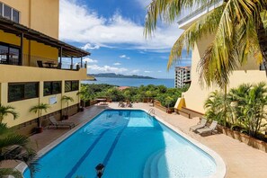 A swimming pool with a view of the sea, surrounded by palm trees and a building with balconies. - Newly Remodeled Flamingo Condo with Sweeping Ocean Views, Spacious Terrace & Walkable Location (Playa Flamingo)