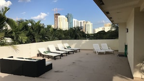 Eine Dachterrasse mit weißen Liegestühlen und einem schwarzen Sofa mit Blick auf eine Skyline mit Hochhäusern und Baukränen.