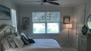 Ein Schlafzimmer mit einem Bett, einem Deckenventilator, einem Fenster mit Jalousien, einer Lampe und einem dekorativen Spiegel.