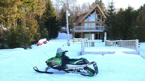Una moto de nieve estacionada frente a una cabaña con terraza de madera y una amplia entrada con ventanas.