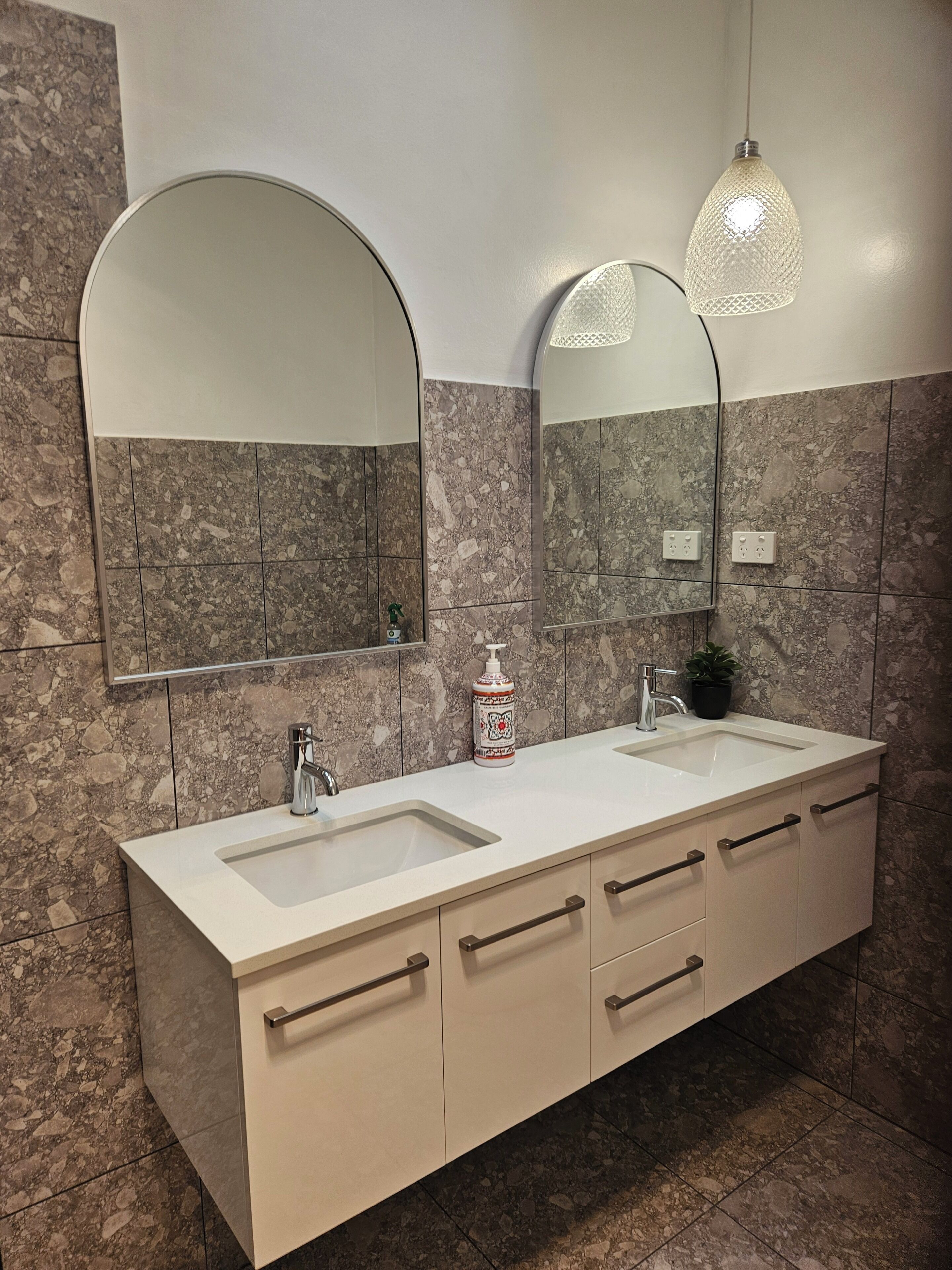 A bathroom with two sinks, a mirror, a faucet, a soap dispenser, and a hanging light fixture.