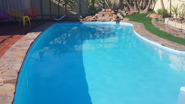 A rectangular swimming pool with a stone border, surrounded by a patio area with chairs and a small rock garden.