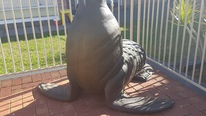 Une grande sculpture représentant un lion de mer, placée sur une surface pavée, avec une clôture métallique en arrière-plan.