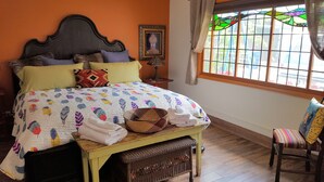 Ein Schlafzimmer mit Bett, Kissen, Nachttisch, einem Korb aus Weidengeflecht, einem Stuhl und einem Fenster mit Ausblick.