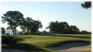 Um campo de golfe com bunkers de areia, um green de aproximação e um corpo de água ao fundo.