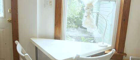 A small room with a white table and two white chairs, a window with a curtain, and a door with a light switch.