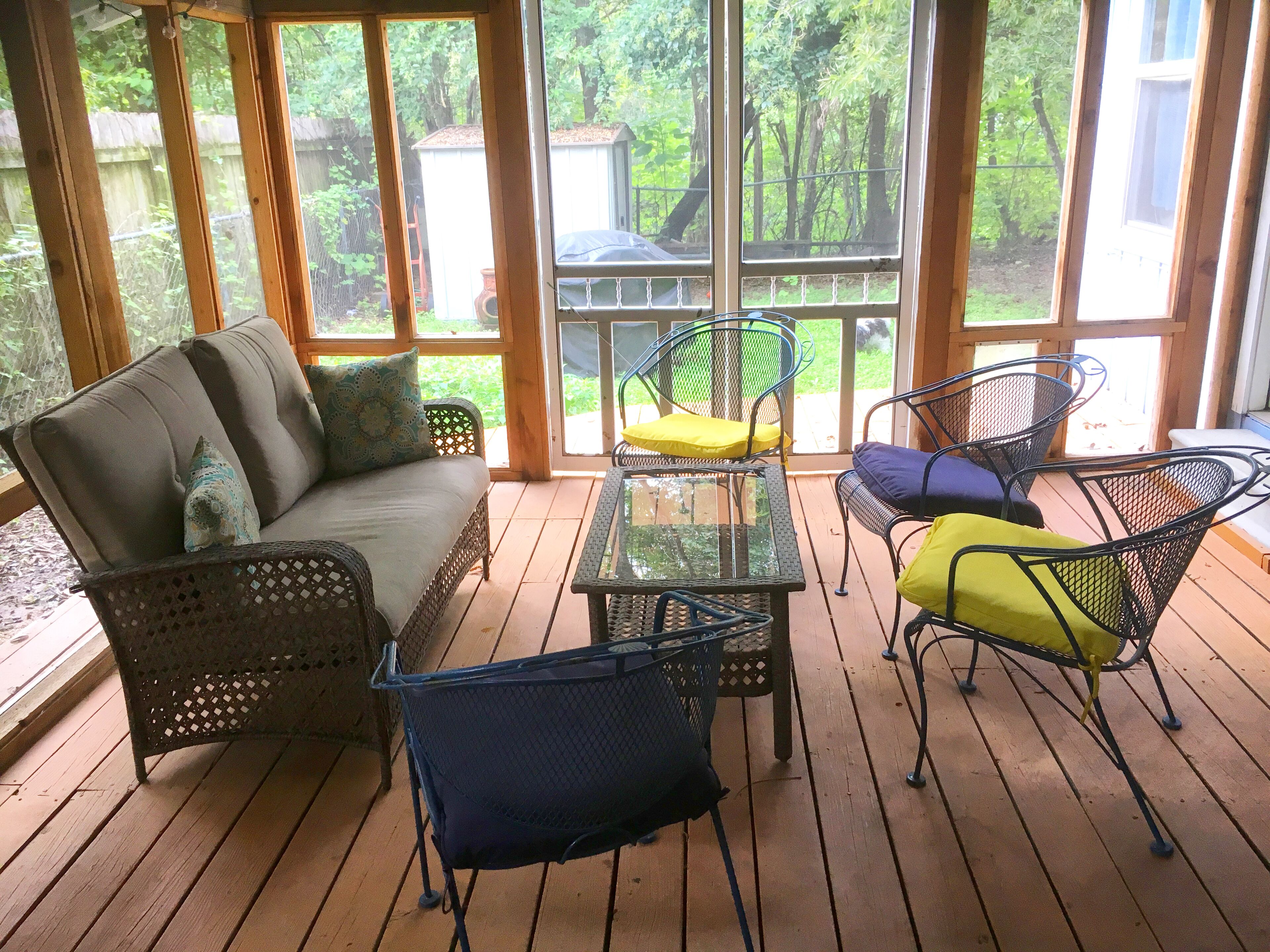 Eine überdachte Terrasse mit Holzdeck, Korbmöbeln und großen Fenstern mit Blick auf den Garten.