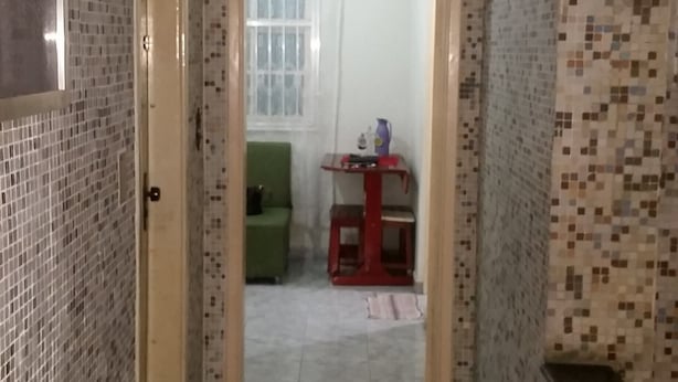 Een smalle gang met betegelde muren, een deur naar een kamer, en een kleine tafel met een vaas en een fles.