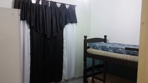 Een kleine kamer met een bed, een houten stoel, een gordijn en een wandplank.