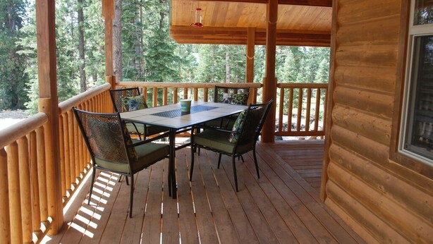 Terraza de madera con mesa y sillas, rodeada por baranda de madera, con vista a un área boscosa.