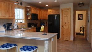 Una cocina con armarios de madera, un refrigerador negro y una encimera de mármol. Hay dos taburetes azules y una ventana con persianas.