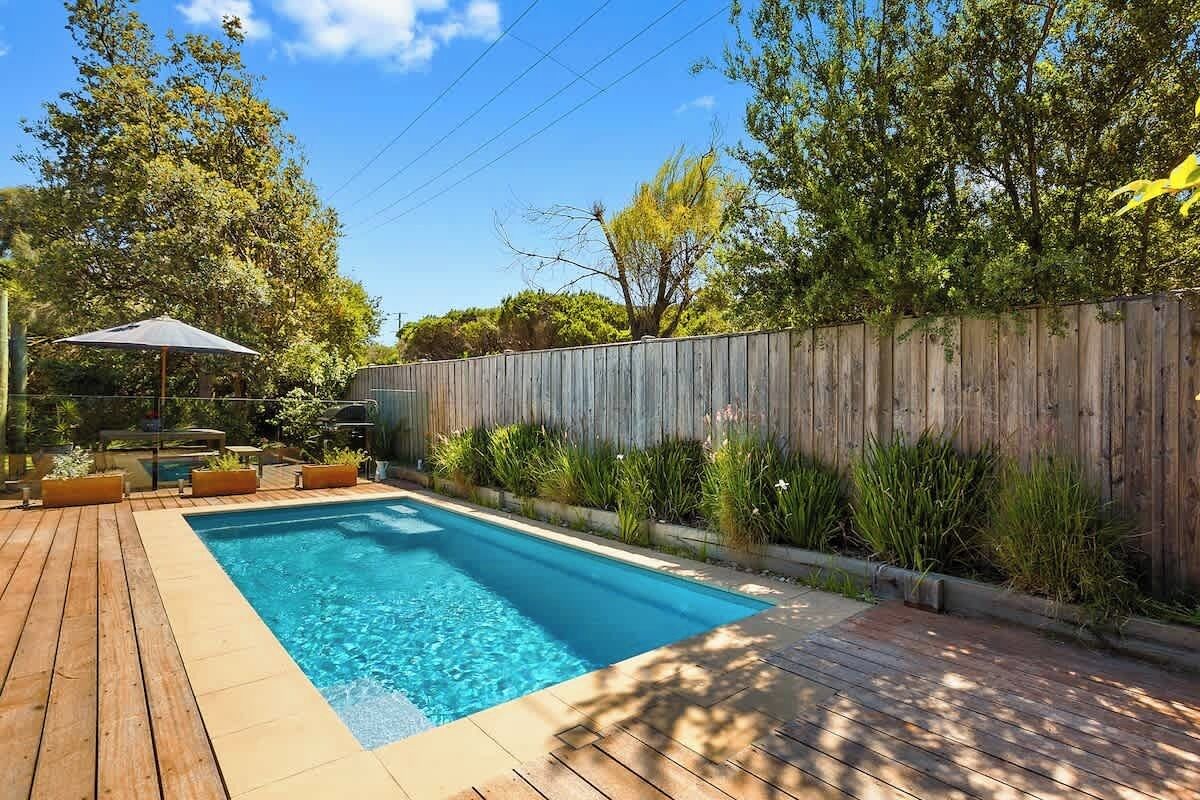A rectangular swimming pool with a wooden deck surrounding it, a wooden fence, and various plants and shrubs.
