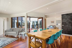 A spacious living area with a wooden dining table, teal chairs, a grey sofa, and a large window.