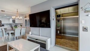 A modern living room with a flat-screen TV, a dining area with a white table and chairs, and a built-in elevator with a control panel. - Ocean View Condo | Pool, Private Beach Access, Elevator, Sleeps 8 (Tybee Island)