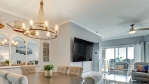 A dining area with a round table, chairs, and a chandelier. There is a television mounted on the wall and a ceiling fan. - Ocean View Condo | Pool, Private Beach Access, Elevator, Sleeps 8 (Tybee Island)