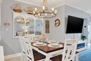 A dining area with a wooden table set for four, white chairs, a chandelier, a large mirror, and a wall-mounted flat-screen TV. - Ocean View Condo on Tybee Island | Pool Access (Tybee Island)