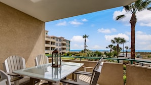 A balcony with a glass table and chairs, overlooking a pool and a building with multiple balconies. - Ocean View Condo | Pool, Private Beach Access, Elevator, Sleeps 8 (Tybee Island)