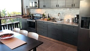 Una cocina moderna con armarios oscuros, un refrigerador de acero inoxidable, horno empotrado y un amplio ventanal con vistas a un jardín.