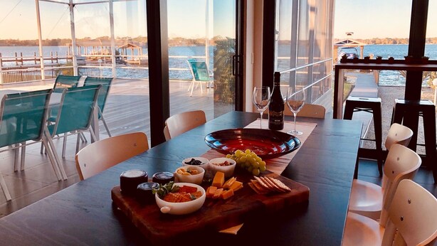 Una mesa de comedor con diversa comida y bebidas, amplias ventanas que ofrecen vistas de un frente marítimo y un puente, y asientos exteriores con sillas.