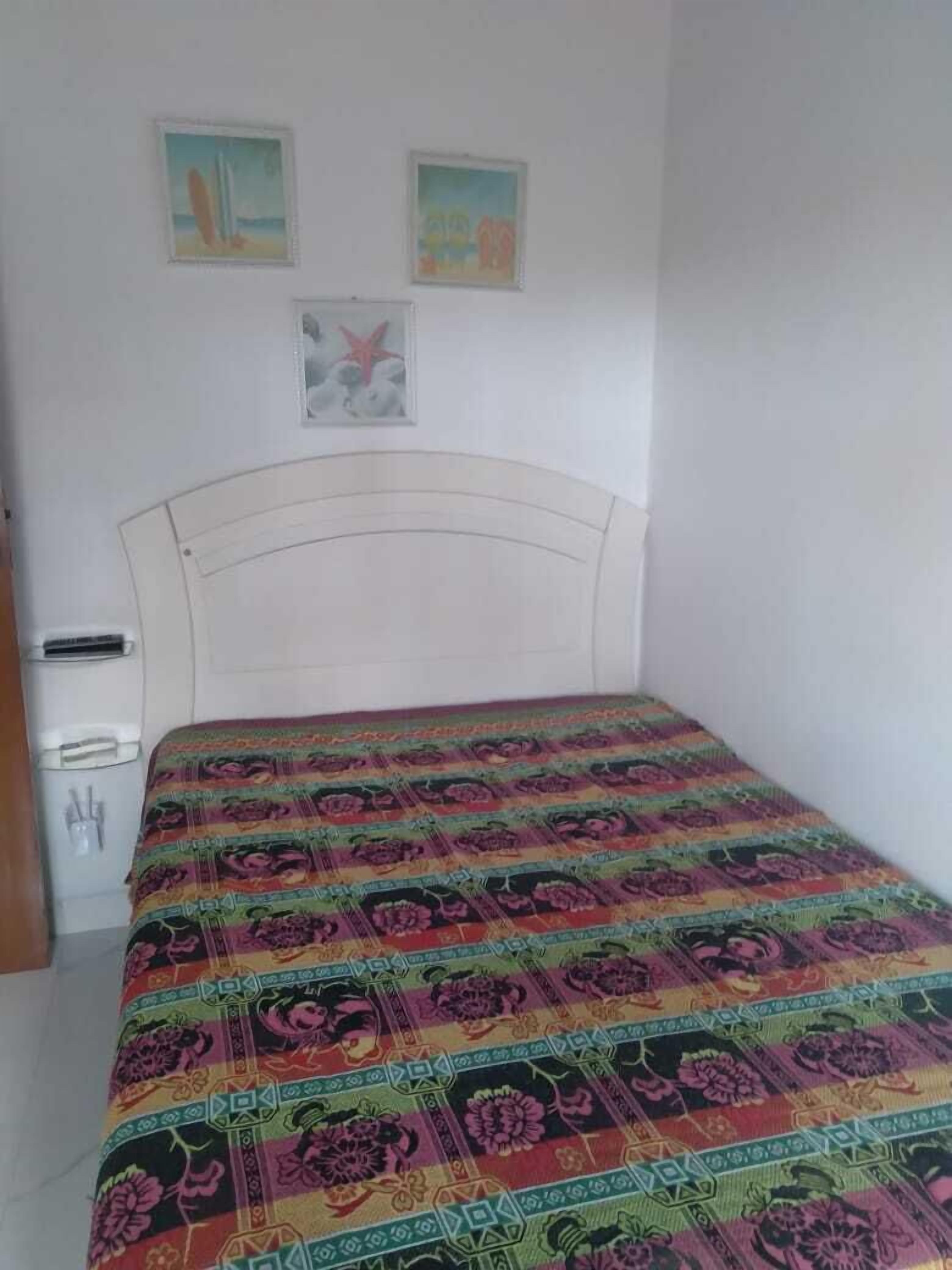A bedroom with a white headboard, a colorful bedspread, a small bedside table, and a wall-mounted heater.