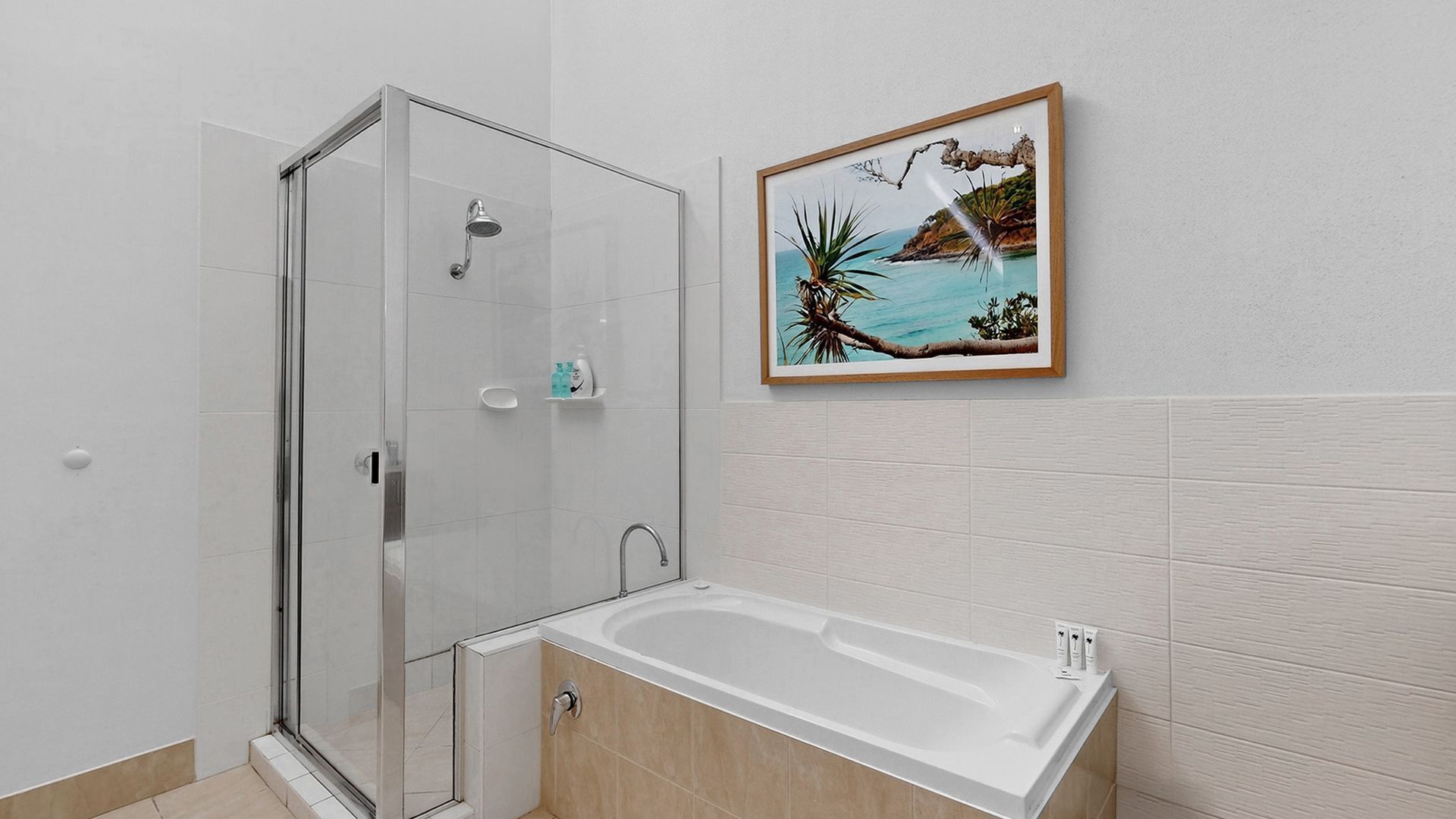 Un bagno moderno con doccia vetrata, una vasca freestanding e un quadro incorniciato raffigurante un paesaggio costiero.