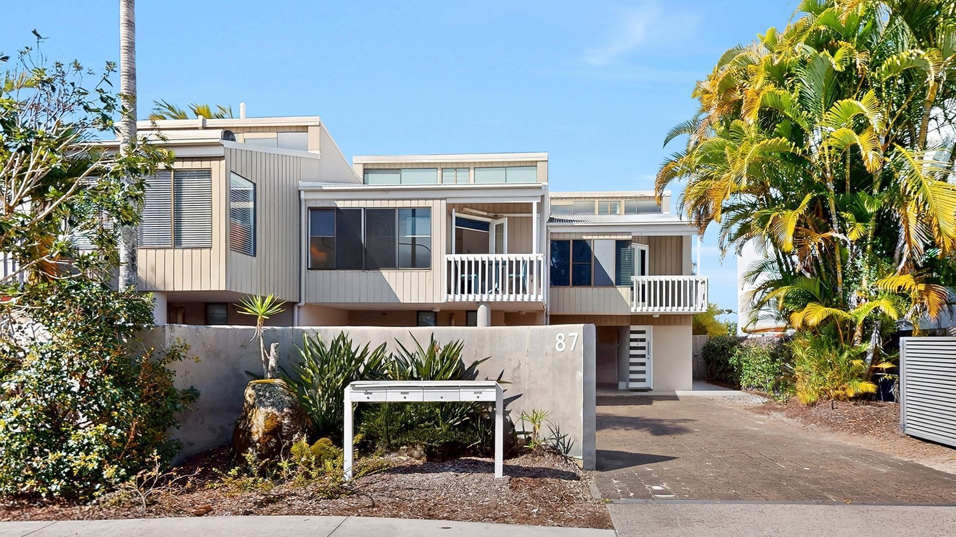 A modern multi-story residential building with balconies, a driveway, and tropical landscaping.