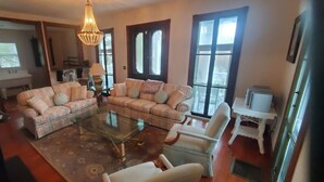 A living room with a glass coffee table, two sofas, and a white armchair. There is a chandelier hanging from the ceiling, and a window with blinds.
