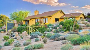 A yellow house with a patio, surrounded by desert vegetation and rocks. - The Joshua Tree Oasis, Joshua Tree Oasis Home (Joshua Tree)