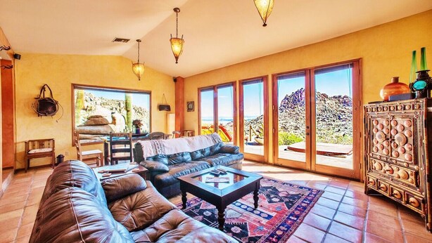 A spacious living room with a leather sofa, a coffee table, and a dining area with chairs. There are large windows offering a view of the outdoors. - The Joshua Tree Oasis, Joshua Tree Oasis Home (Joshua Tree)