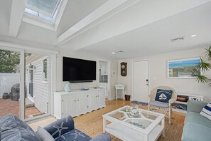 A spacious living room with a flat-screen TV, a white cabinet, a wicker chair, and a view of the outdoors. - Cape Escape - Walk to Beach, Fenced Yard, A/C & Pickleball (South Chatham)