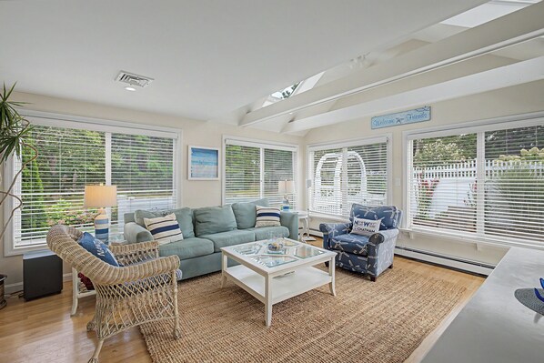 A spacious living room with a light blue sofa, a wicker chair, a glass coffee table, and a blue armchair with nautical patterns. - Cape Escape - Walk to Beach, Fenced Yard, A/C & Pickleball (South Chatham)
